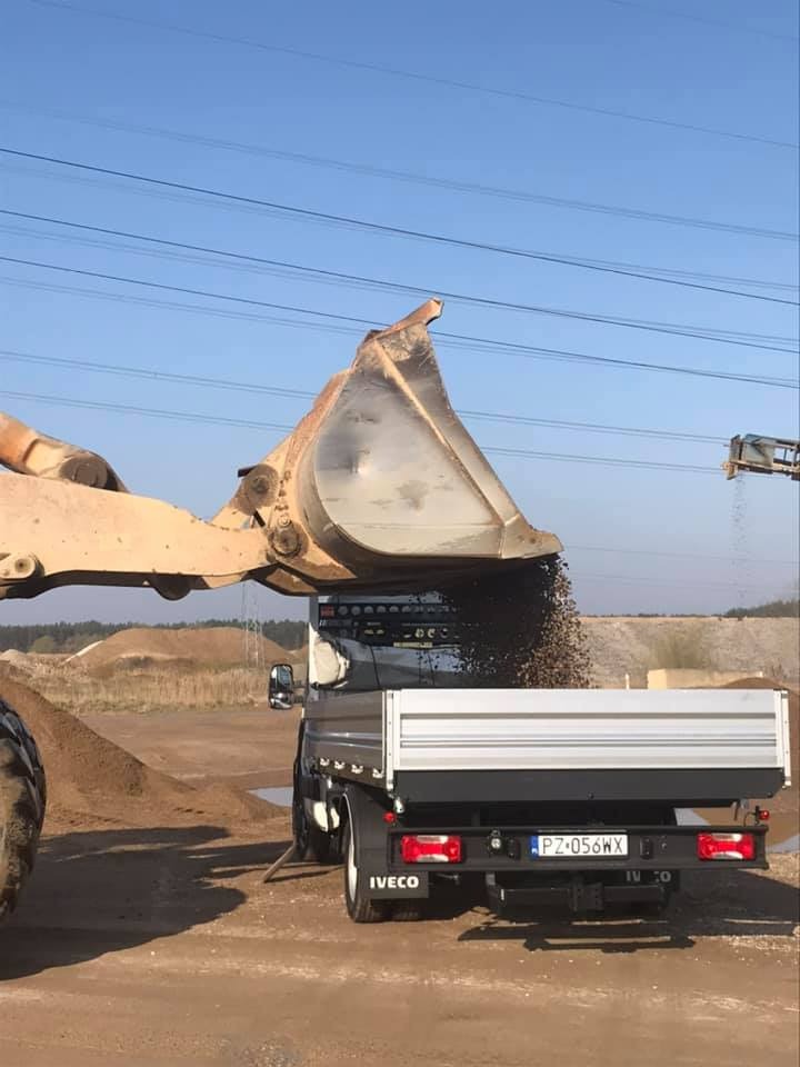 Ładowarka czołowa z łyżką wysypuje żwir do skrzyni ładunkowej czarnego busa Iveco na tle hałd ziemi i błękitnego nieba.