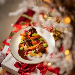 Wykwintne danie mięsne z puree, marchewką, groszkiem i sosem, udekorowane jadalnymi kwiatami, podane na białym talerzu, umieszczonym na stosie prezentów świątecznych, z rozmytymi lampkami...