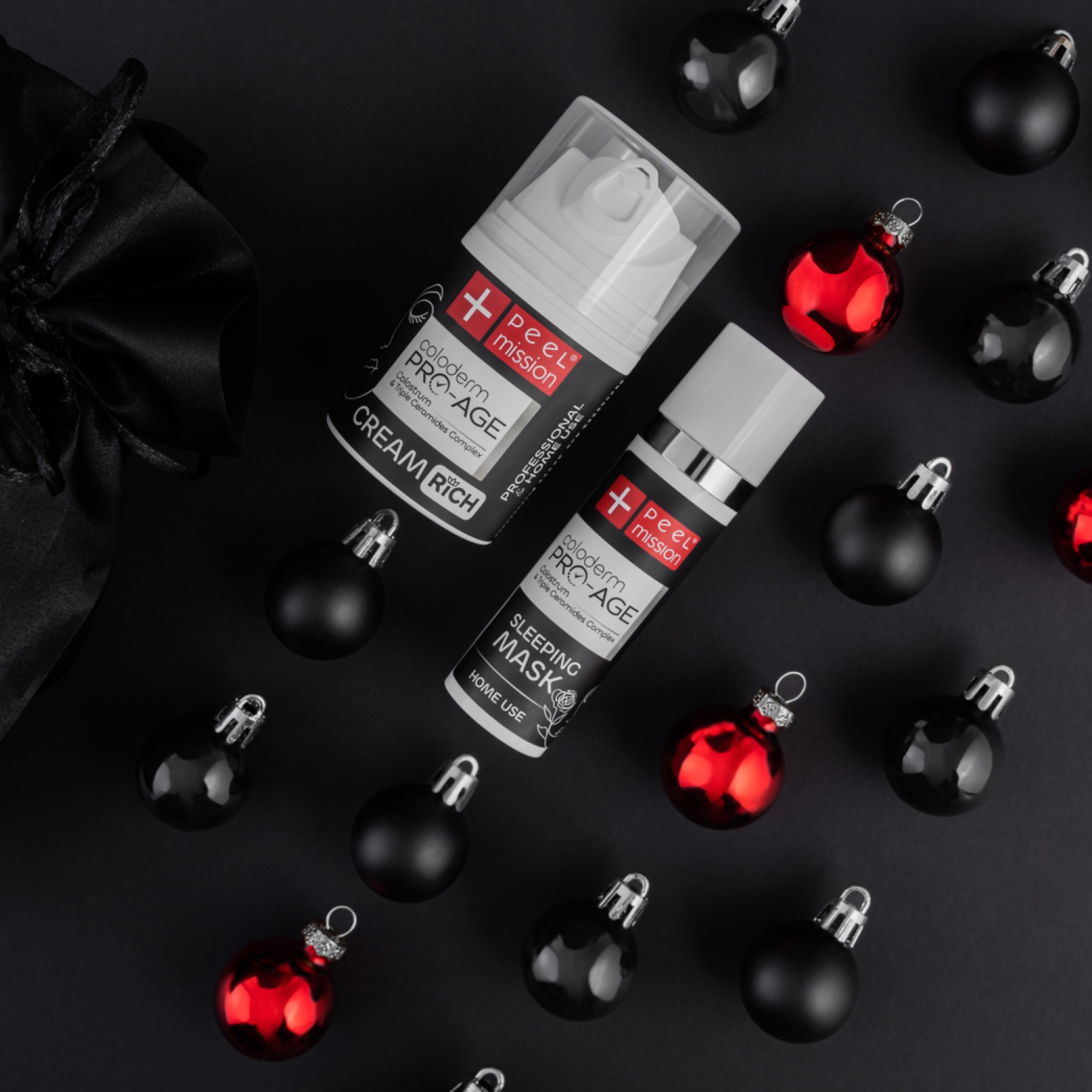 Dwa opakowania kosmetyków Coloderm Pro-Age, krem bogaty i maska na noc, otoczone czarnymi i czerwonymi bombkami na czarnym tle, obok czarny woreczek ze wstążką.
