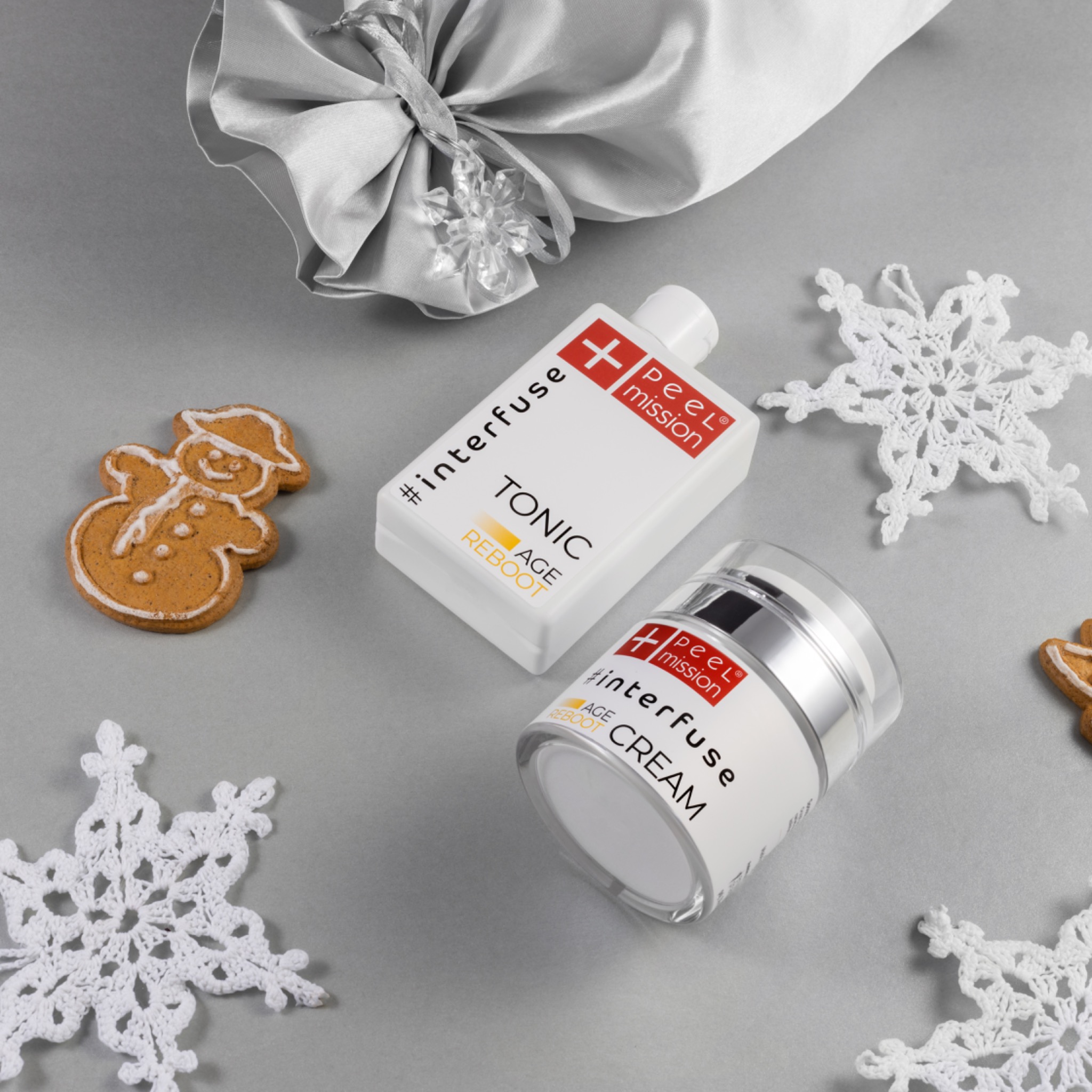 Zestaw kosmetyków: tonik i krem do twarzy marki Interfuse Peel Mission, otoczone pierniczkiem w kształcie bałwana i dekoracjami w kształcie płatków śniegu, całość umieszczona na srebrnym tle...