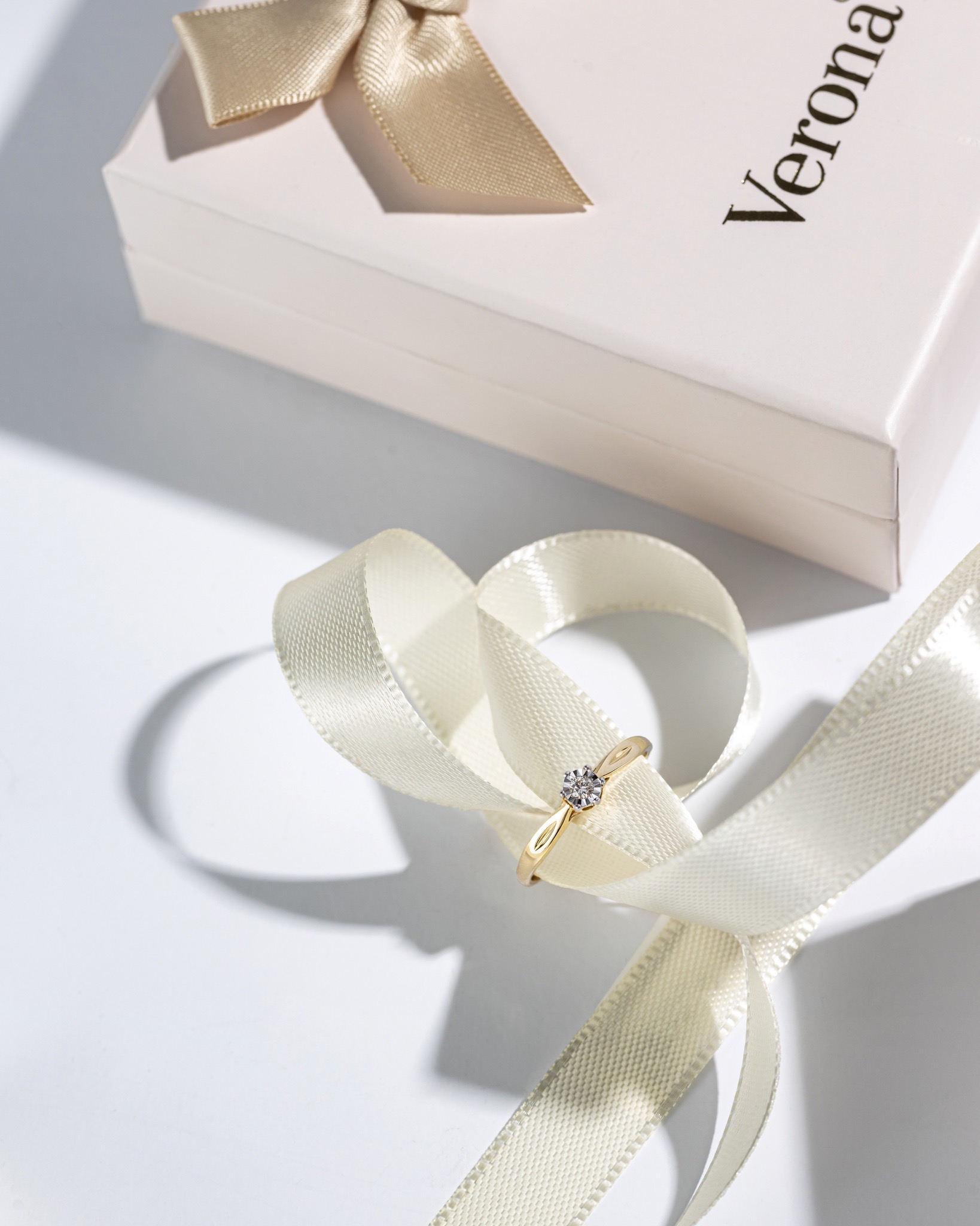 Złoty pierścionek z diamentem na kremowej wstążce, obok pudełko z logo Verona, na białym tle w jasnym oświetleniu.