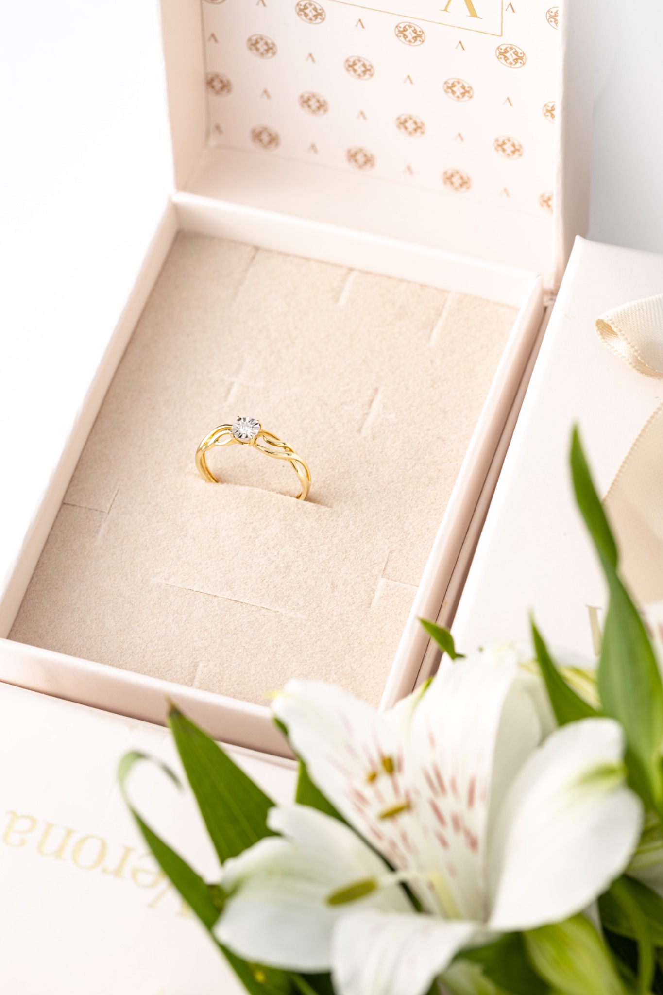 Złoty pierścionek z pojedynczym brylantem w otwartym, jasnoróżowym pudełku z kwiatami alstroemeria w rogu.