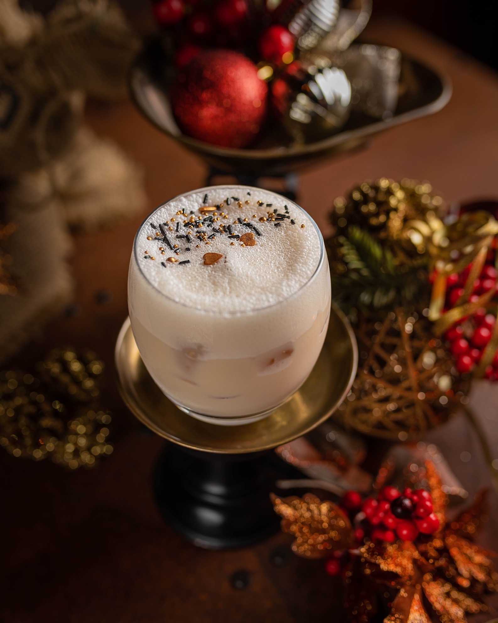 Kremowy koktajl w szklance na złotej podstawce, posypany czarno-złotymi kuleczkami, otoczony świątecznymi dekoracjami w odcieniach złota i czerwieni.