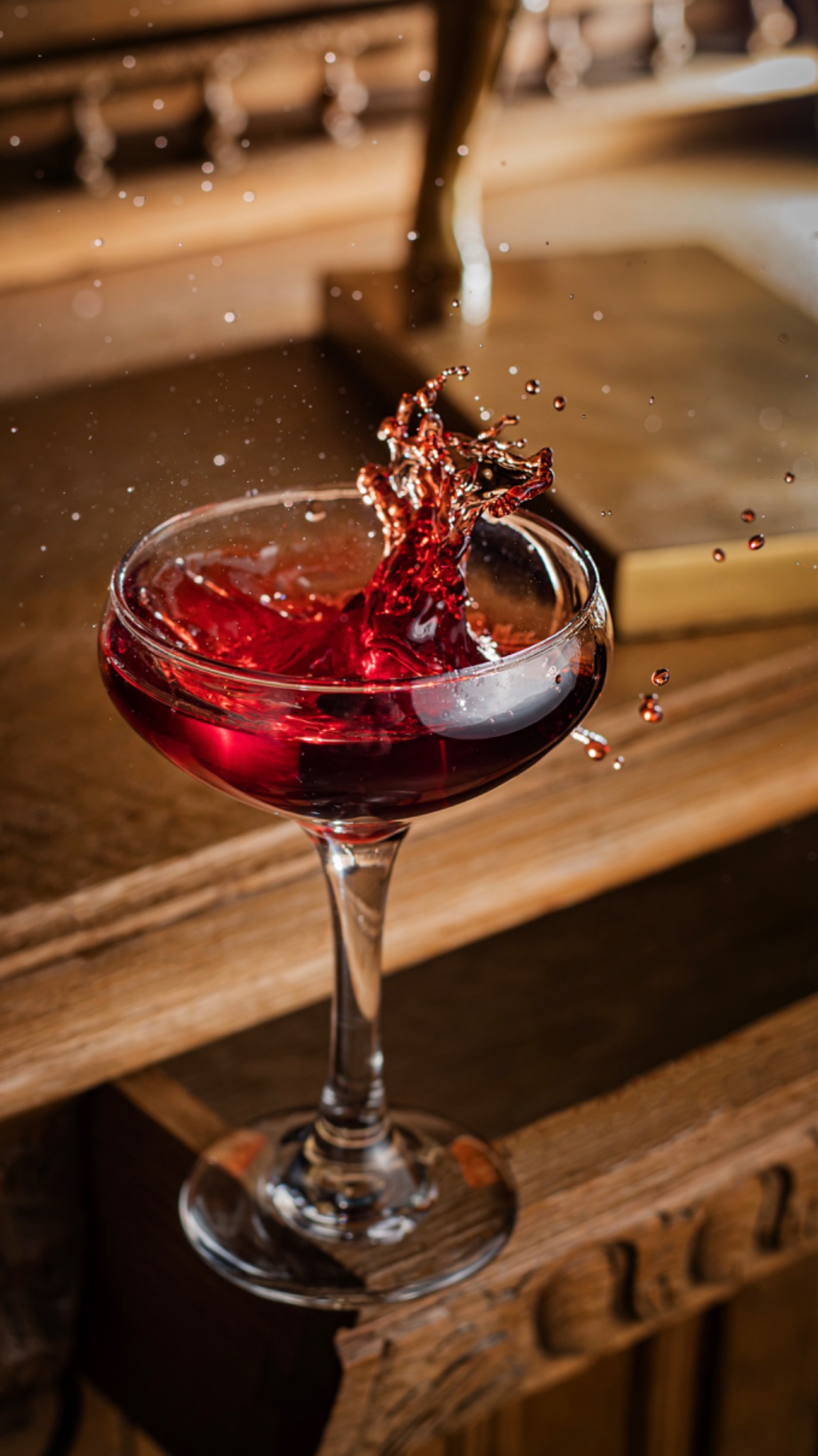 Kieliszek z czerwonym płynem, w którym widać moment rozprysku, umieszczony na rzeźbionym, drewnianym stole, w tle fragment drewnianej kolumny.