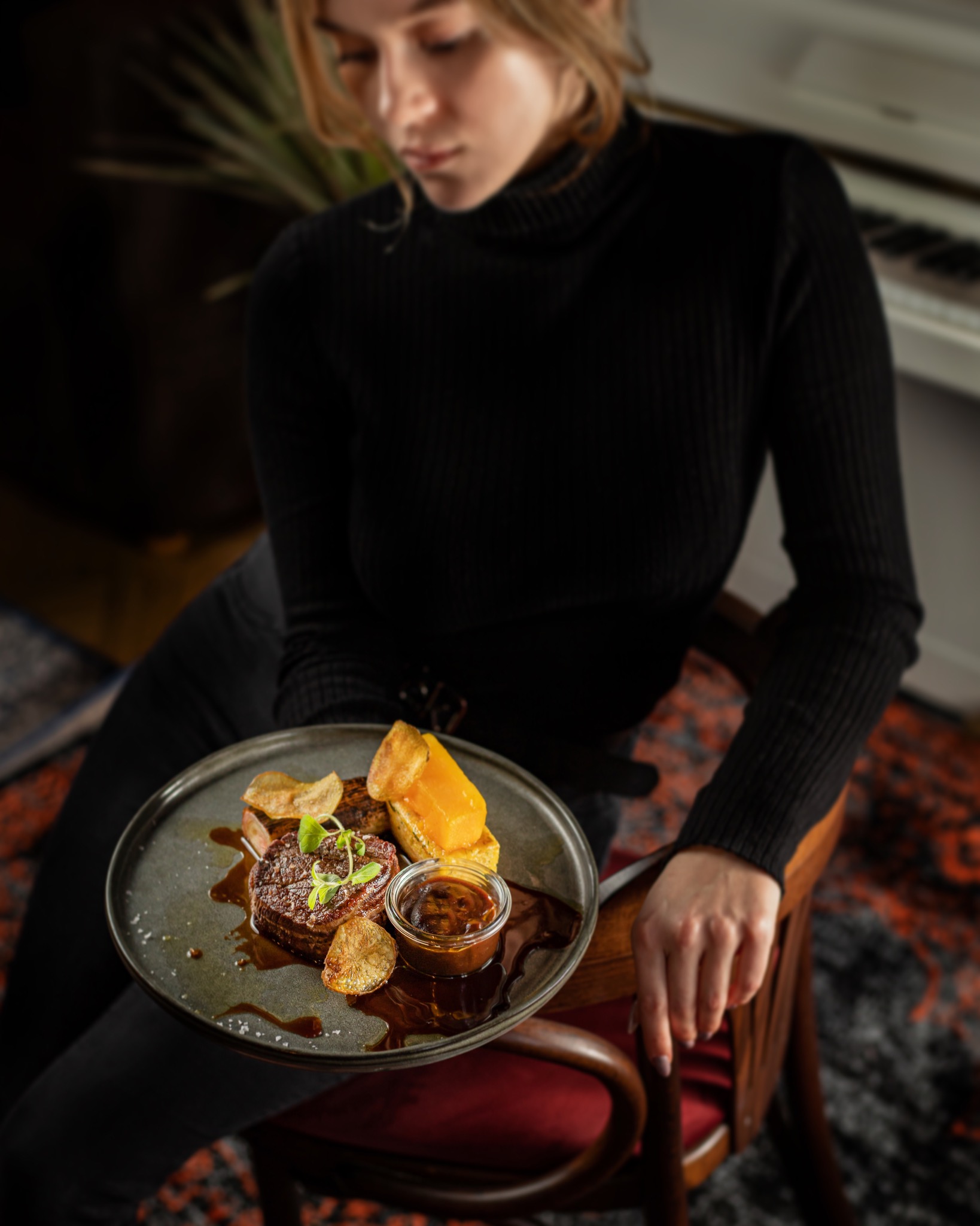 Kobieta siedzi na krześle i trzyma talerz z wyszukanym daniem: stek, sos w słoiczku, chipsy i kawałek sera.