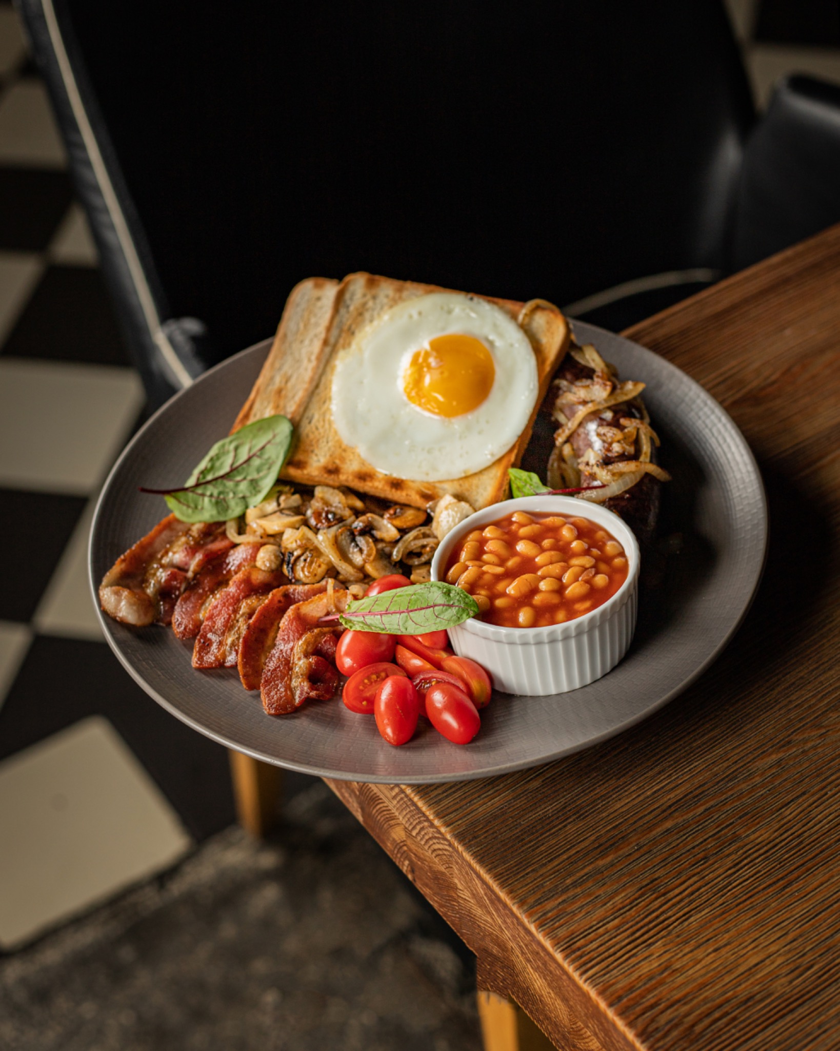 Pełny angielski śniadaniowy zestaw na szarym talerzu: jajko sadzone na tostach, bekon, pieczarki, pomidorki cherry, fasolka w sosie pomidorowym w białej miseczce, całość udekorowana zielonymi...