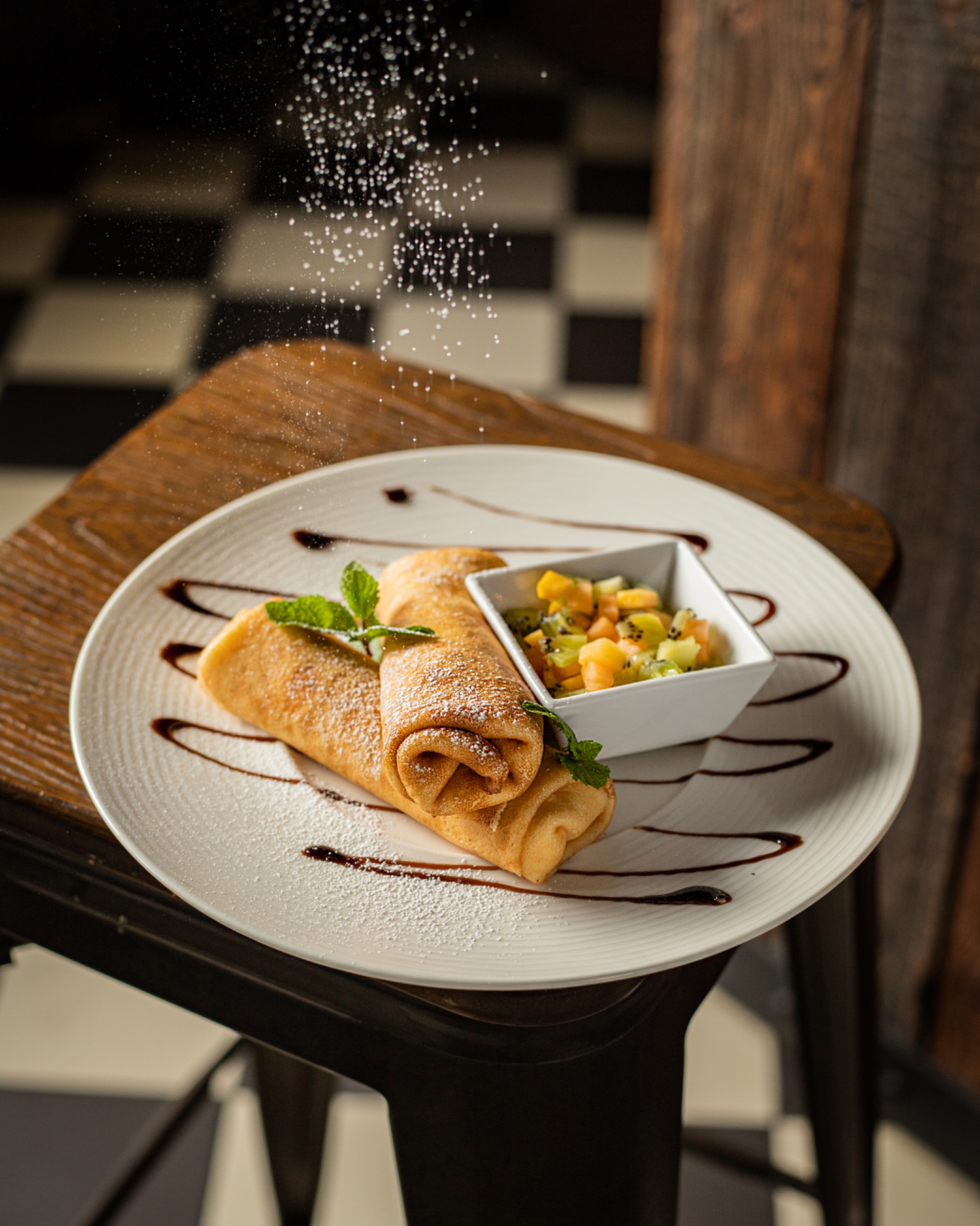 Dwa naleśniki posypane cukrem pudrem, udekorowane miętą i sosem czekoladowym, podane z sałatką owocową w białej miseczce na białym talerzu, na tle krzesła i ściany w szachownicę.