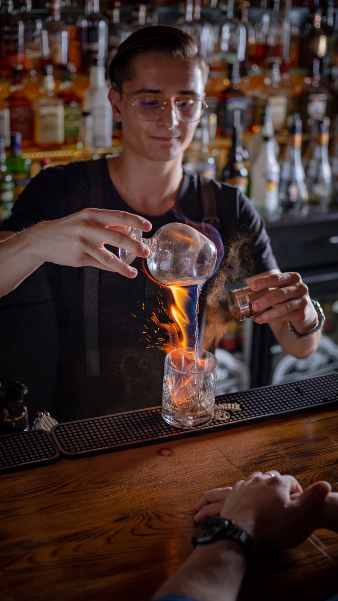 Barman w okularach przygotowuje płonący koktajl, nalewając płyn z karafki i posypując przyprawami do szklanki z lodem, na tle półek z butelkami w barze.