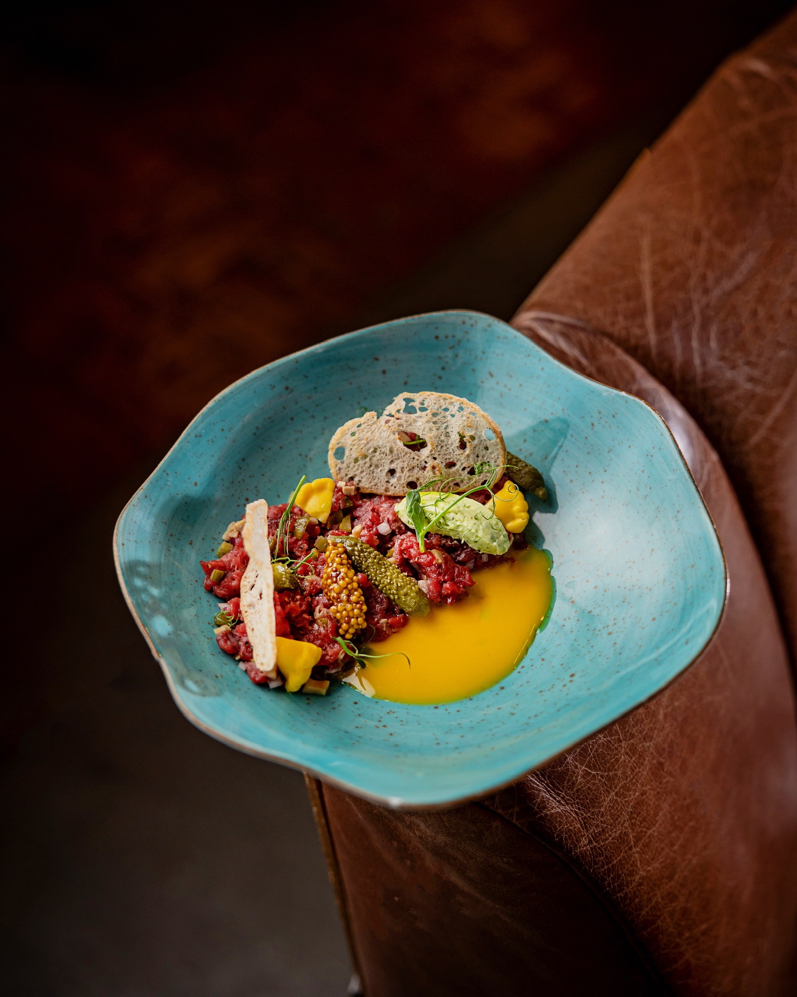 Tatar wołowy na turkusowym talerzu, z żółtkiem, grzankami i marynatami, ułożony na skórzanym fotelu w restauracji.