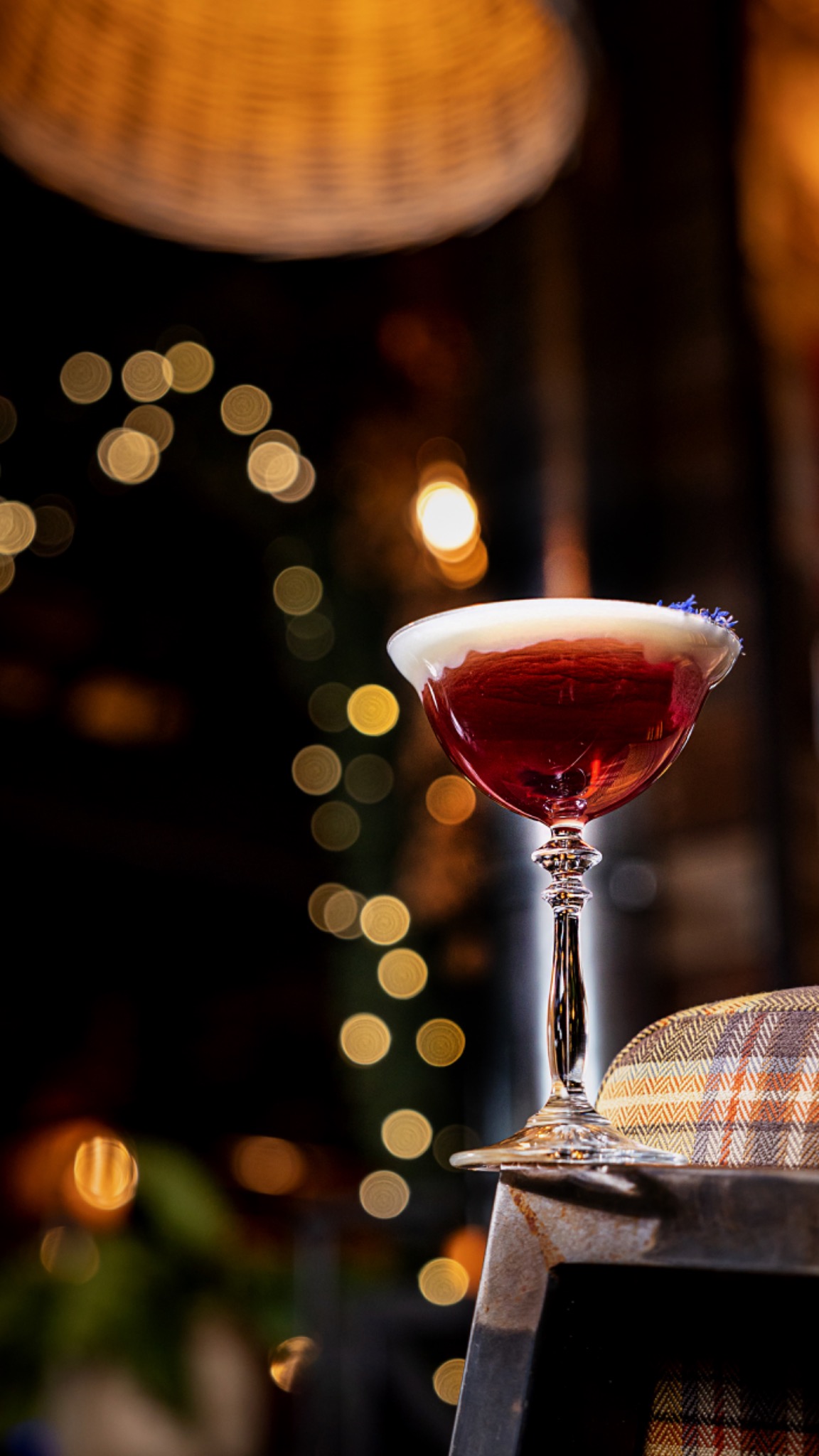 Elegancki kieliszek z czerwonym koktajlem zwieńczonym białą pianką i drobnymi niebieskimi kwiatami, ustawiony na metalowym stoliku obok wzorzystej czapki; w tle rozmyte światła.