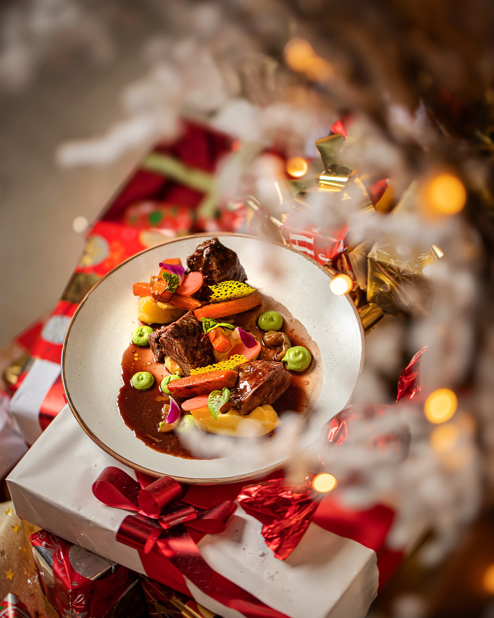 Wykwintne danie mięsne z puree, marchewką, groszkiem i sosem, udekorowane jadalnymi kwiatami, podane na białym talerzu, umieszczonym na stosie prezentów świątecznych, z rozmytymi lampkami...