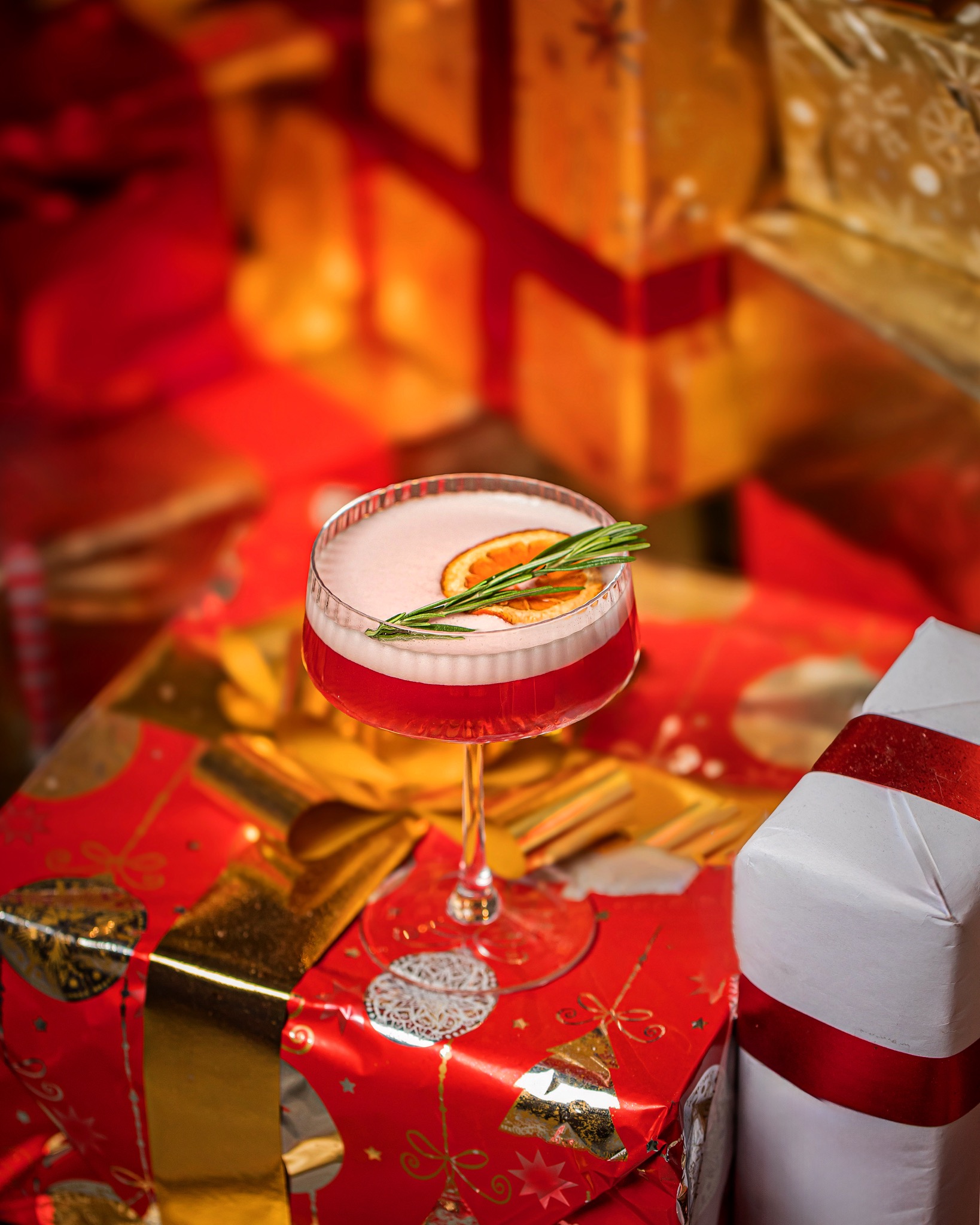 Czerwony koktajl z pianką, plastrem pomarańczy i gałązką rozmarynu, umieszczony na błyszczących, świątecznie zapakowanych prezentach.