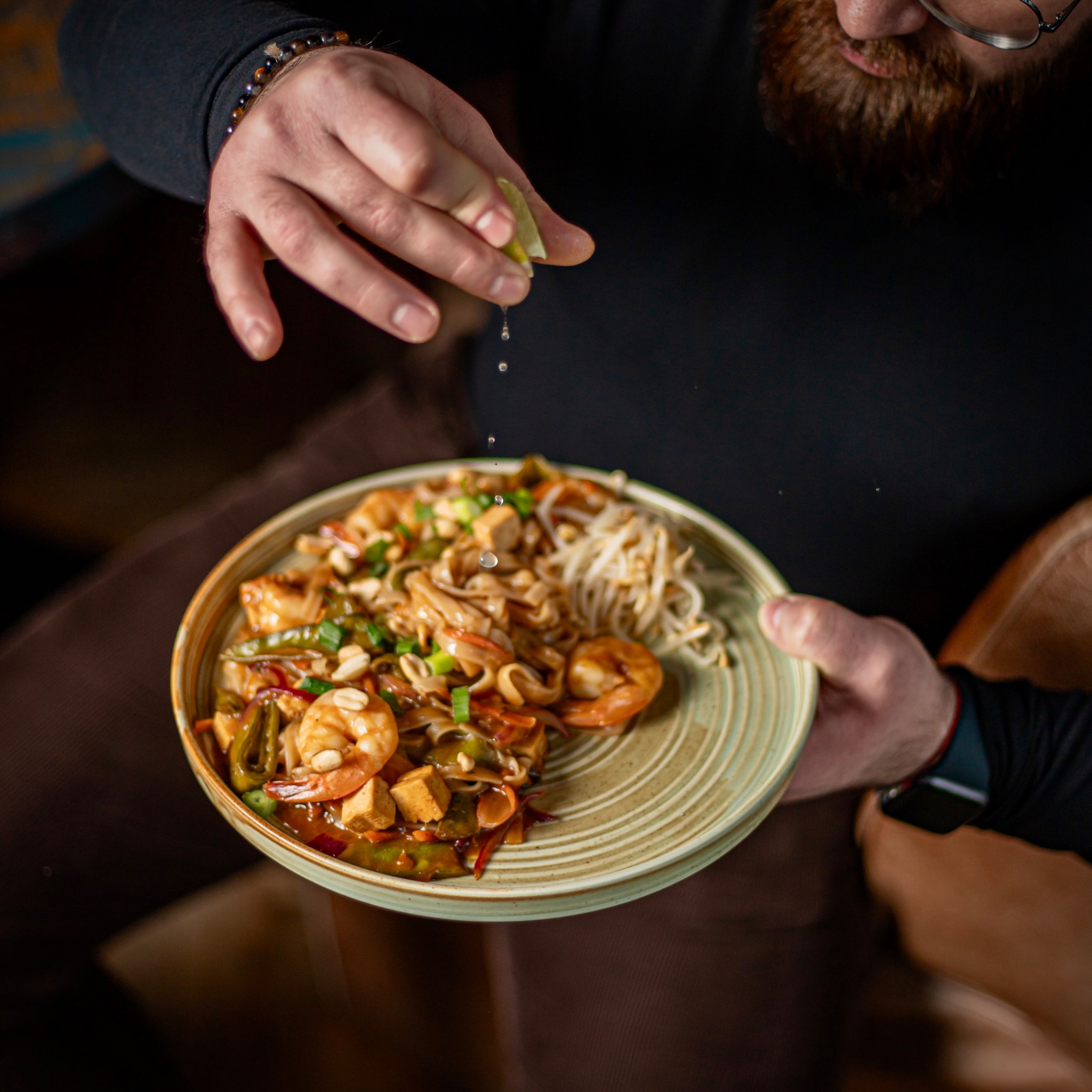 Kucharz wyciska limonkę na pad thai z krewetkami, tofu i orzechami na talerzu z zielonymi paskami.
