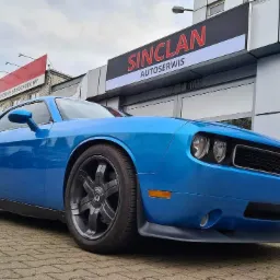 Błękitny samochód marki Dodge Challenger zaparkowany przed budynkiem serwisu samochodowego Sinclan Autoserwis, widok z niskiej perspektywy.
