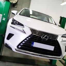 Sinclan Sp. z o.o. Sp. k. - Biały Lexus na podnośniku samochodowym w warsztacie, widok z przodu na atrapę chłodnicy i reflektory.
