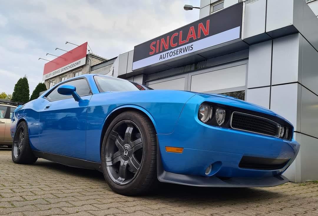 Błękitny samochód marki Dodge Challenger zaparkowany przed budynkiem serwisu samochodowego Sinclan Autoserwis, widok z niskiej perspektywy.