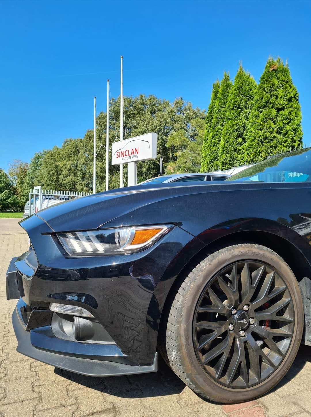 Częściowy widok czarnego Forda Mustanga z przodu, z widocznym logo warsztatu Sinclan Autoservis w tle, na tle zieleni i błękitnego nieba.