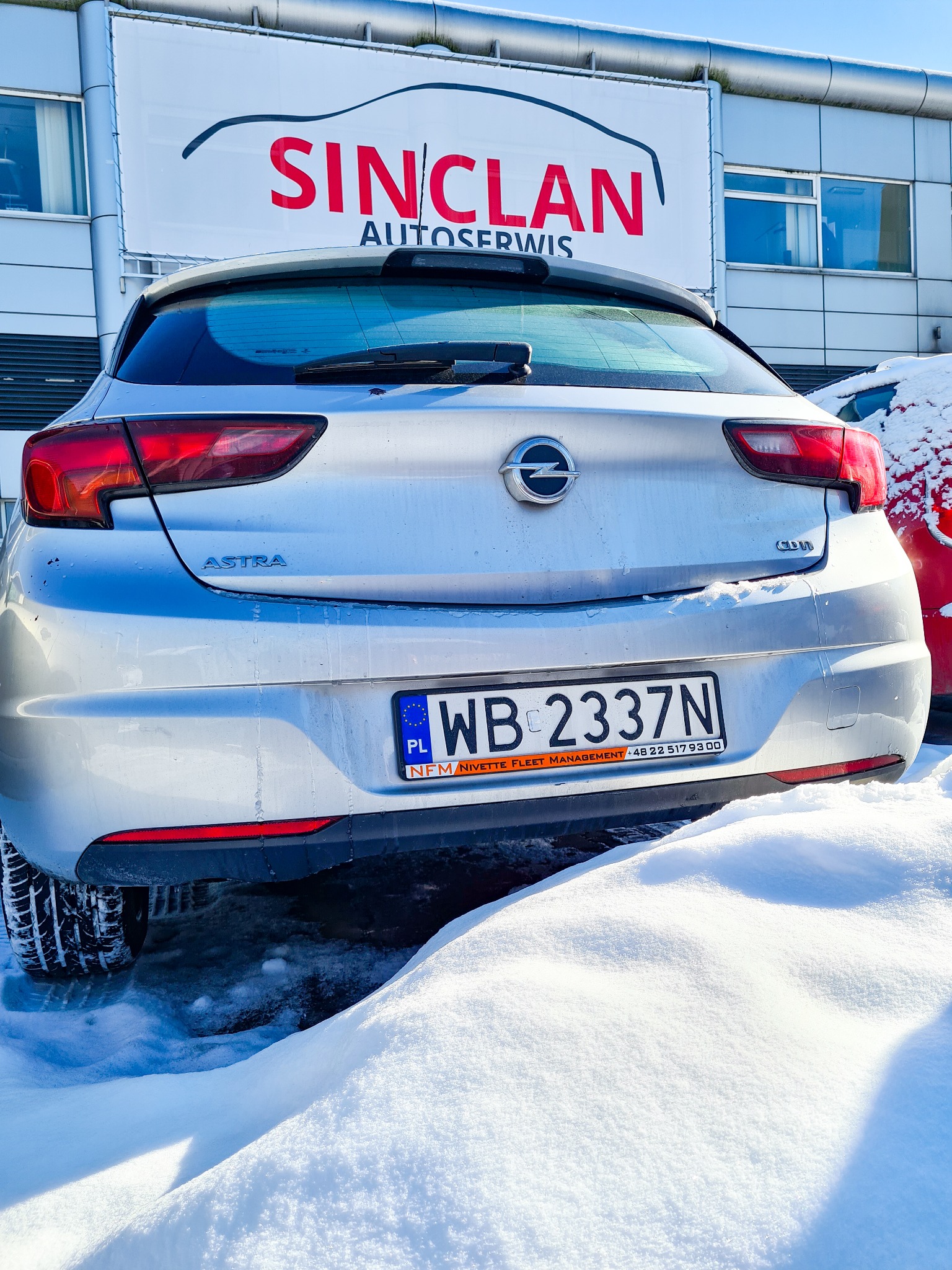 Srebrna Opel Astra stoi zaparkowana na śniegu przed budynkiem z szyldem 'SINCLAN Autoservis'.