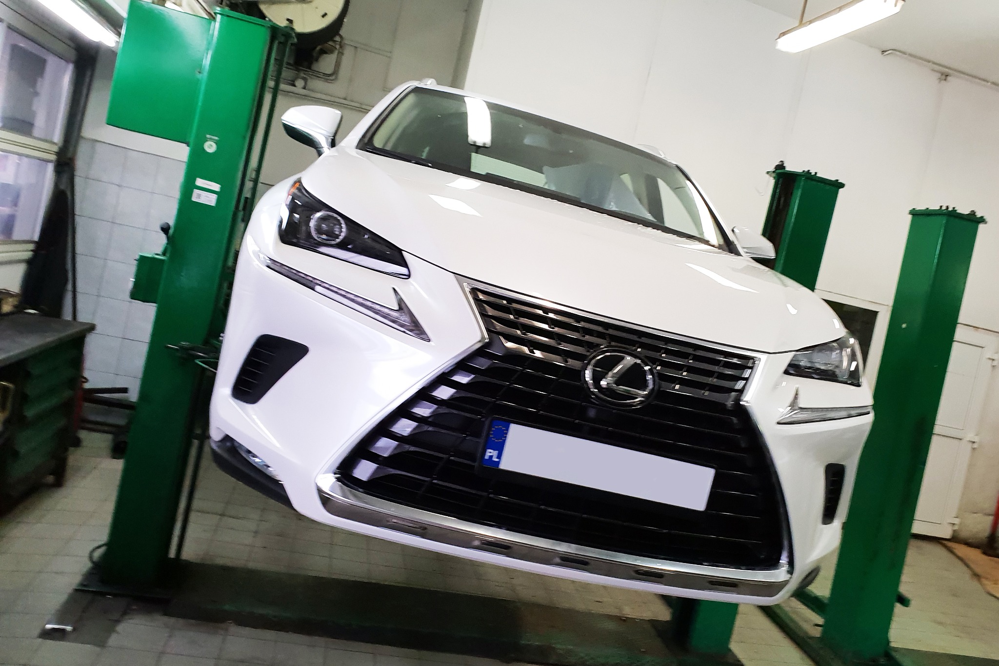 Biały Lexus na podnośniku samochodowym w warsztacie, widok z przodu na atrapę chłodnicy i reflektory.