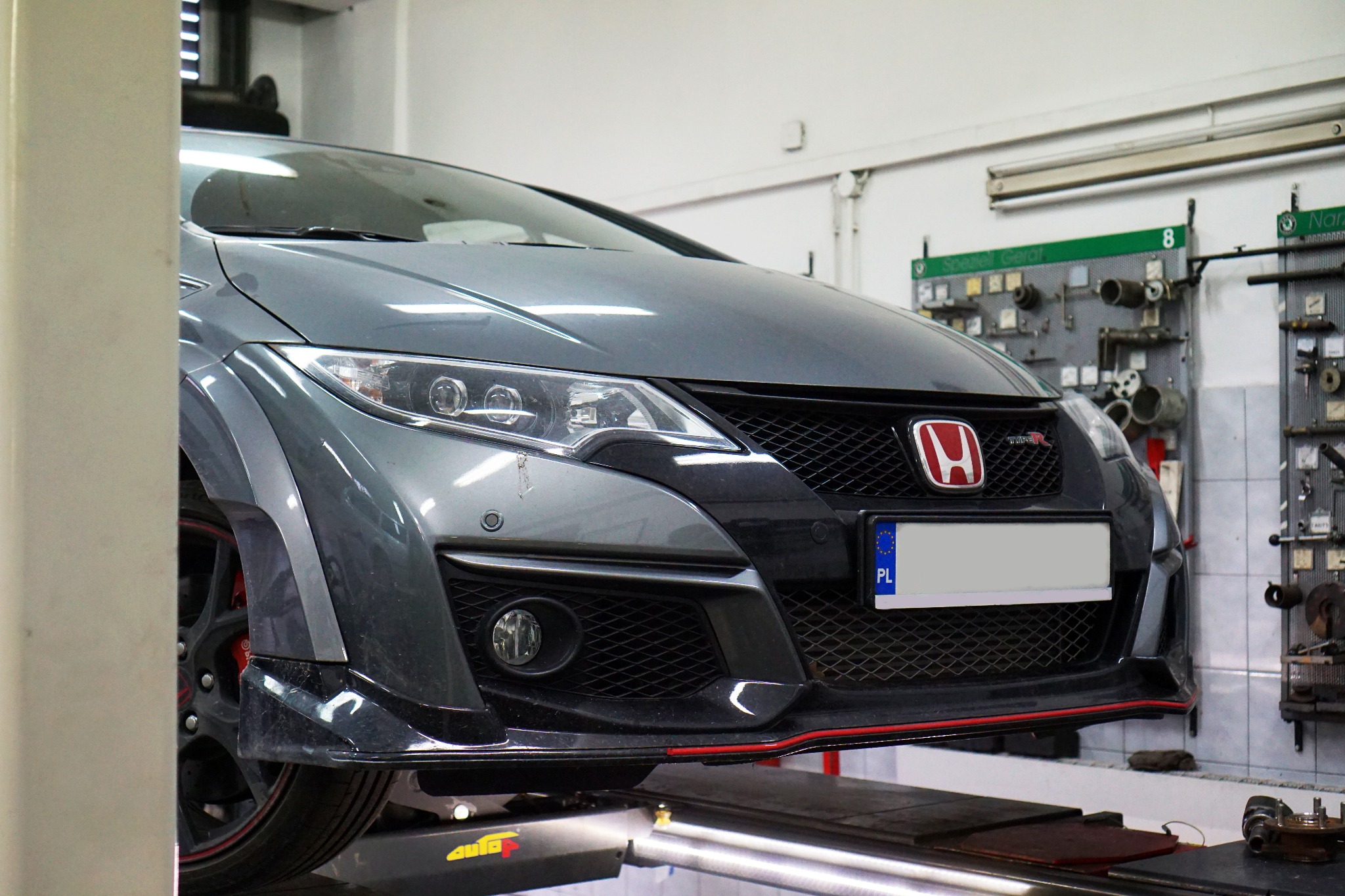 Szary samochód Honda Civic Type R na podnośniku warsztatowym, widoczny przód pojazdu, reflektory, grill z czerwonym logo, tablica rejestracyjna PL, w tle ściana warsztatu z narzędziami.