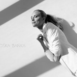 Agnes Modelska - Portret kobiety w białym garniturze, opierającej się o ścianę w ostrym świetle, z widocznym logo 'GOŚKA BAŃKA'.