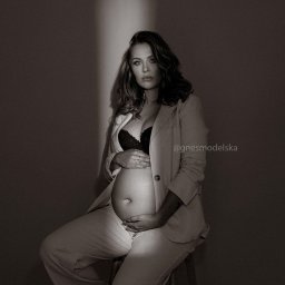 Agnes Modelska - Czarno-białe zdjęcie kobiety w ciąży, siedzącej na drewnianym stołku, ubranej w jasny garnitur i czarny biustonosz, z rękami na brzuchu, oświetlonej pionową smugą światła.