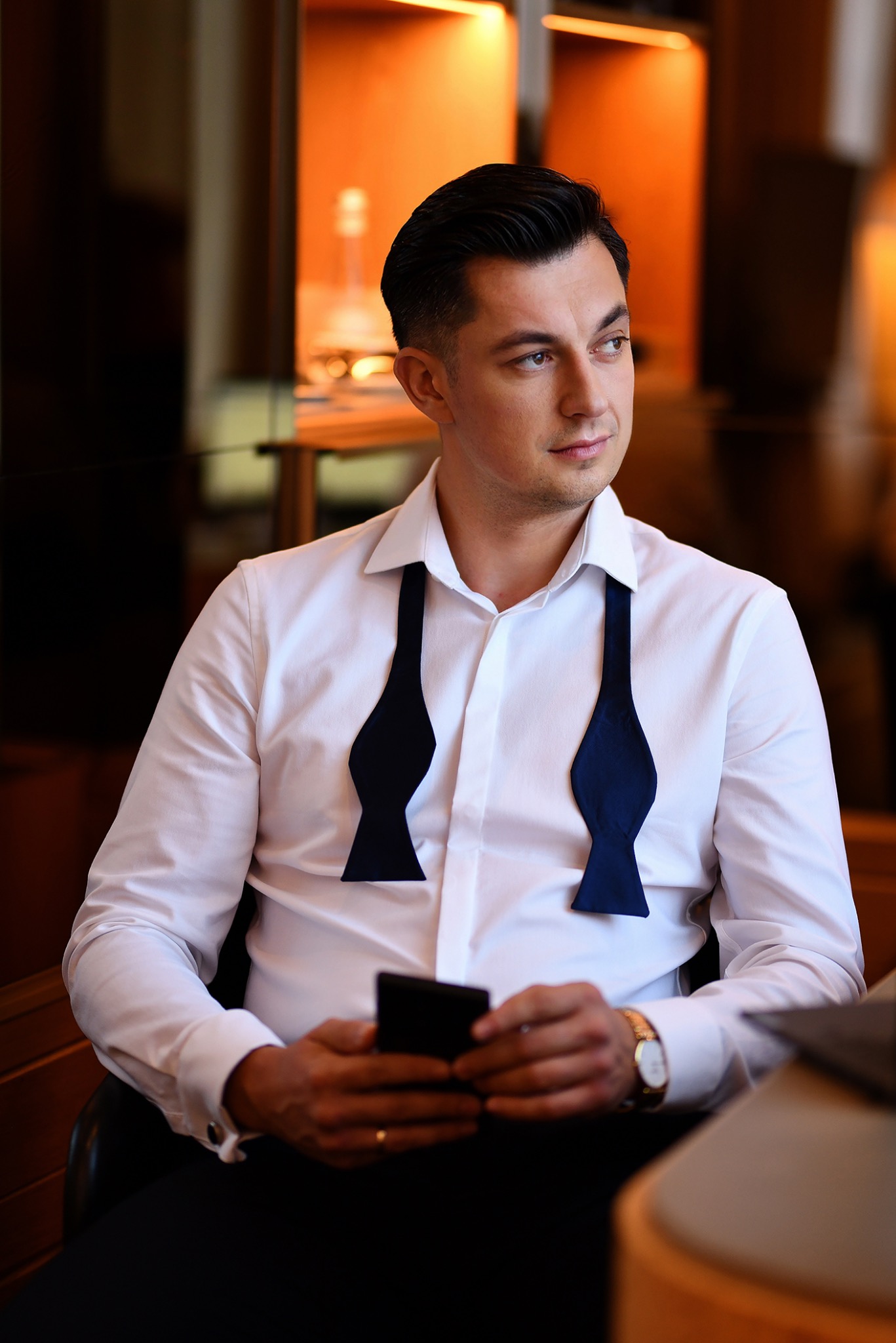 Elegancki mężczyzna w rozpiętej białej koszuli z granatową muszką, trzymający smartfon, z zegarkiem na nadgarstku, spoglądający w bok.