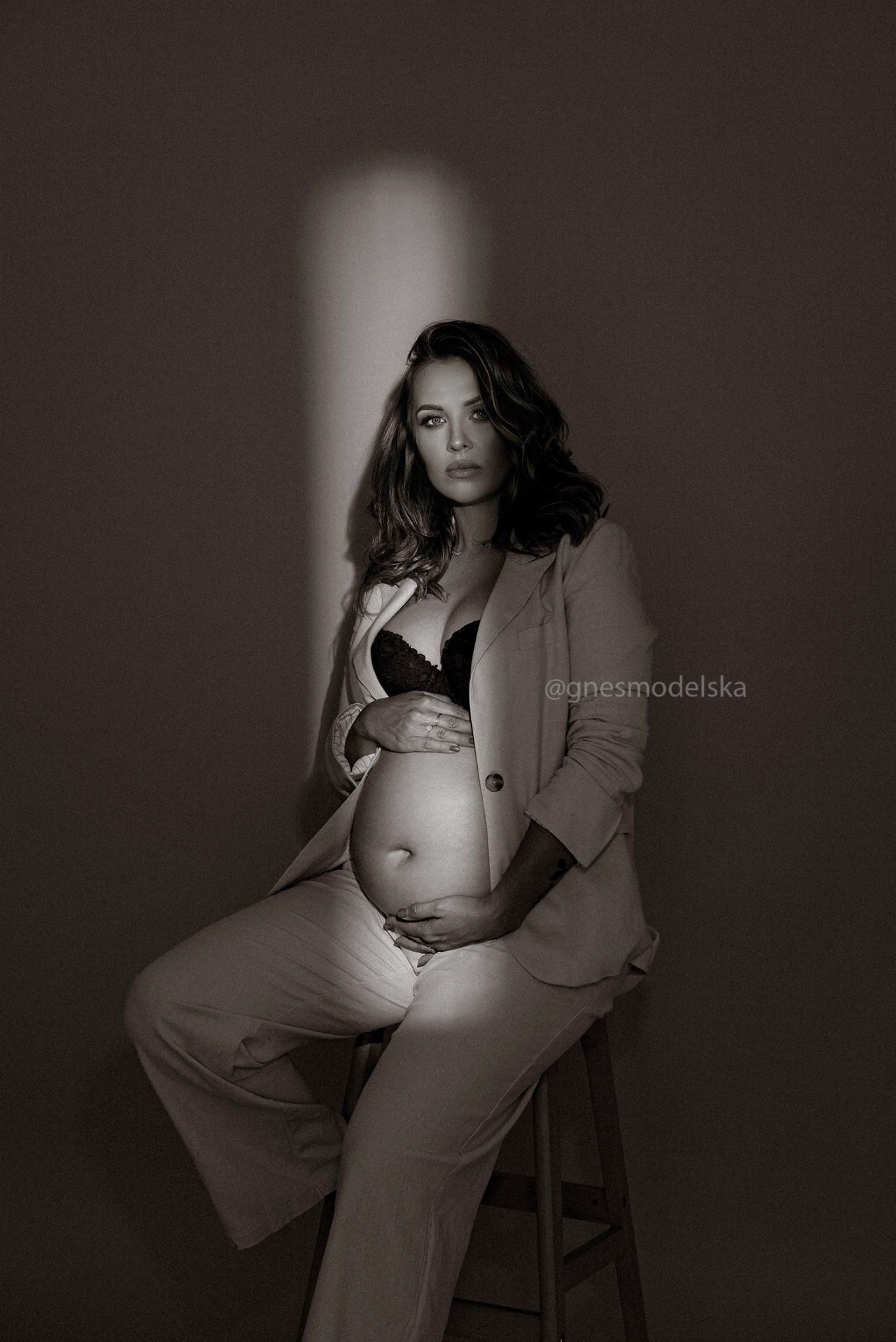 Czarno-białe zdjęcie kobiety w ciąży, siedzącej na drewnianym stołku, ubranej w jasny garnitur i czarny biustonosz, z rękami na brzuchu, oświetlonej pionową smugą światła.