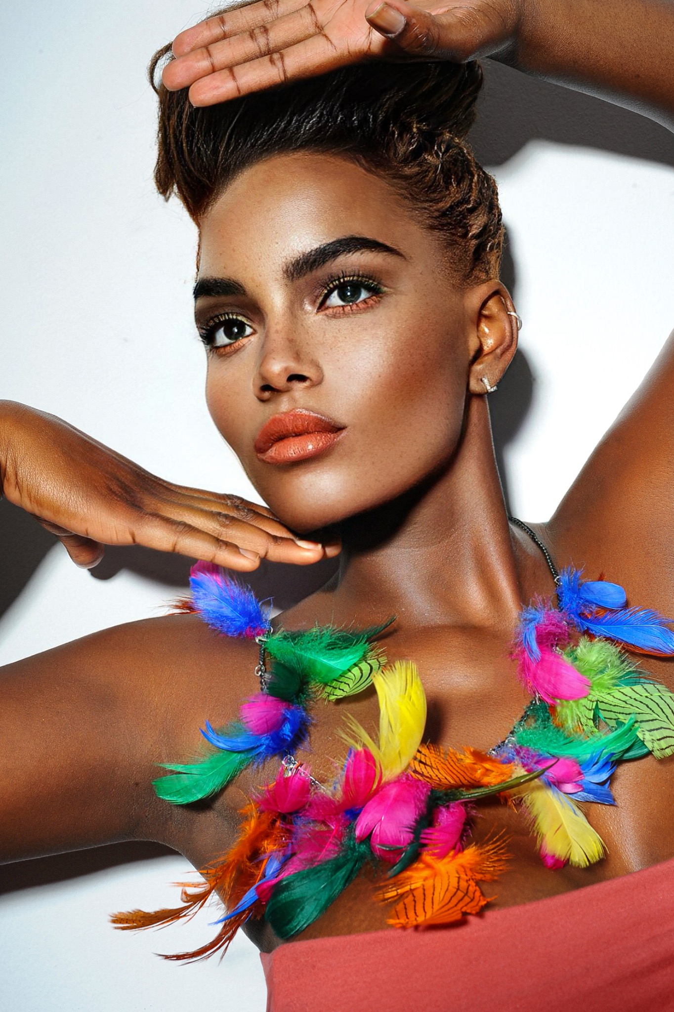 Portret kobiety z ciemną karnacją, w ekstrawaganckim naszyjniku z kolorowych piór, pozującej z ręką uniesioną nad głową i dłonią pod brodą, na jasnym tle.