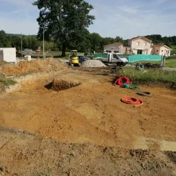 Wykonany wykop pod fundamenty domu jednorodzinnego, widoczne rury instalacyjne, mała koparka i ciężarówka z materiałami budowlanymi w tle.
