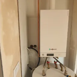 Biały piec gazowy marki Viessmann zamontowany nad białym zasobnikiem ciepłej wody, widoczne miedziane rury instalacji, czarne węże i zawory z czerwoną rączką.