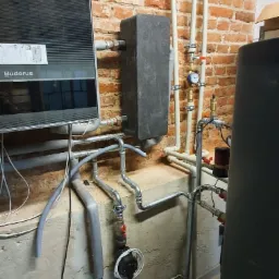 Zamontowany piec Buderus z szarą obudową, zbiornik wyr&oacute;wnawczy, rury instalacji grzewczej z manometrem na tle ściany z czerwonej cegły.