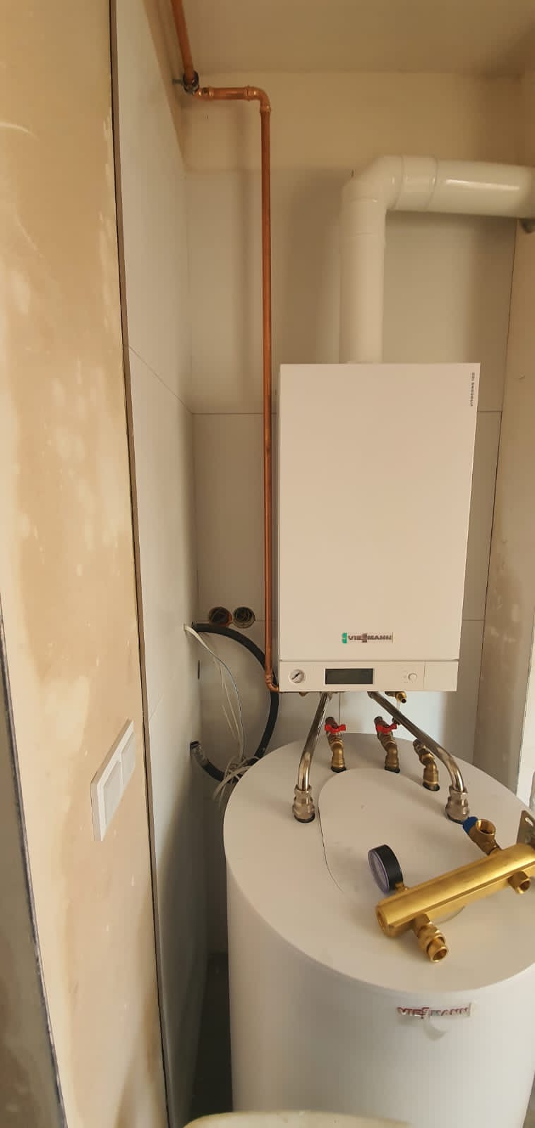 Biały piec gazowy marki Viessmann zamontowany nad białym zasobnikiem ciepłej wody, widoczne miedziane rury instalacji, czarne węże i zawory z czerwoną rączką.