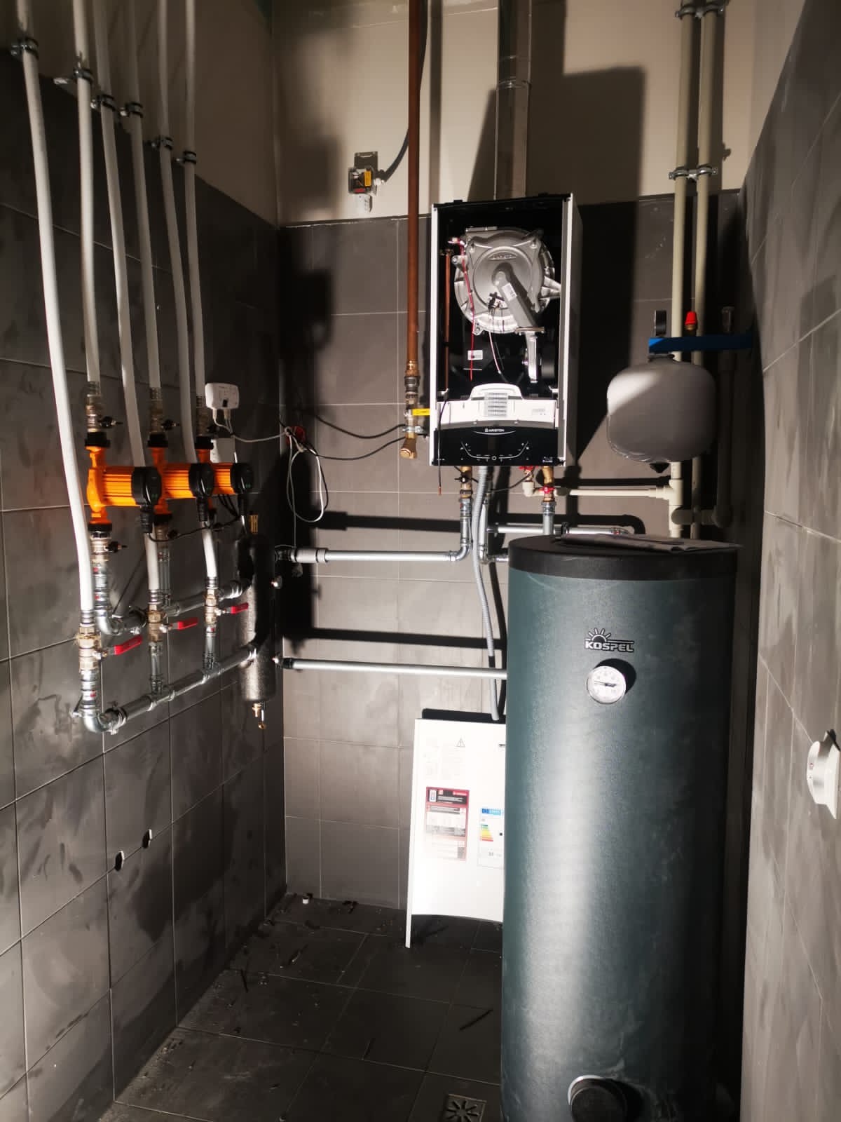 Nowoczesna kotłownia z piecem gazowym, zasobnikiem wody marki Kospel, pompą obiegową z pomarańczową obudową i licznymi rurami, zamontowana w pomieszczeniu wyłożonym szarymi płytkami.