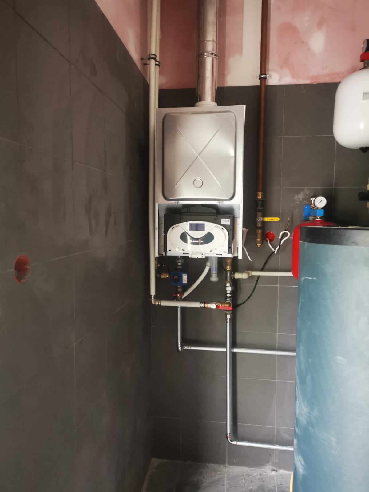 Nowy, wiszący piec gazowy Ariston z podłączonymi rurami, zamontowany na szarej, kafelkowej ścianie, obok zbiornika akumulacyjnego w pomieszczeniu w trakcie remontu.
