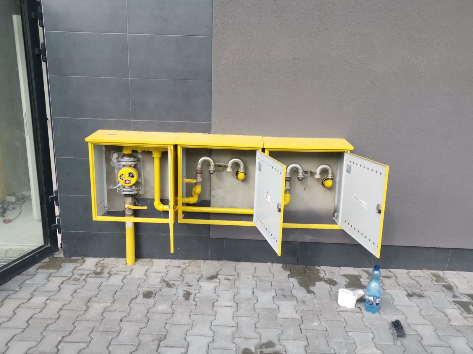 Żółta skrzynka gazowa z otwartymi drzwiczkami, widoczne zawory i rury, butelka wody i kubek na kostce brukowej.