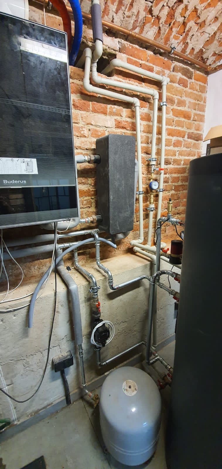 Zamontowany piec Buderus z szarą obudową, zbiornik wyrównawczy, rury instalacji grzewczej z manometrem na tle ściany z czerwonej cegły.