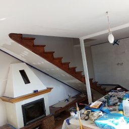 KrzyśBud - Przygotowania do prac remontowych w salonie: zabezpieczone elementy taśmą malarską, zakryte meble, widoczne narzędzia i materiały malarskie, schody drewniane, kominek z ceglaną podstawą.
