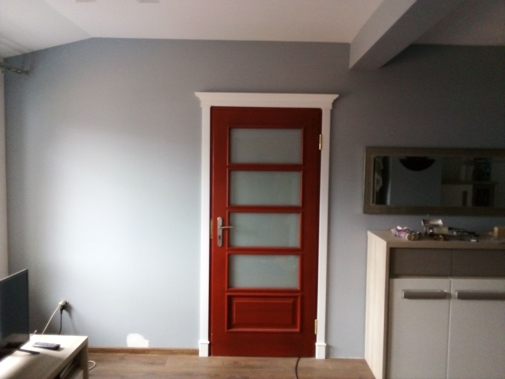 Pomalowane na szaro ściany pomieszczenia z zamontowanymi ciemnoczerwonymi drzwiami z czterema szybami, białe listwy przypodłogowe i ościeżnica.