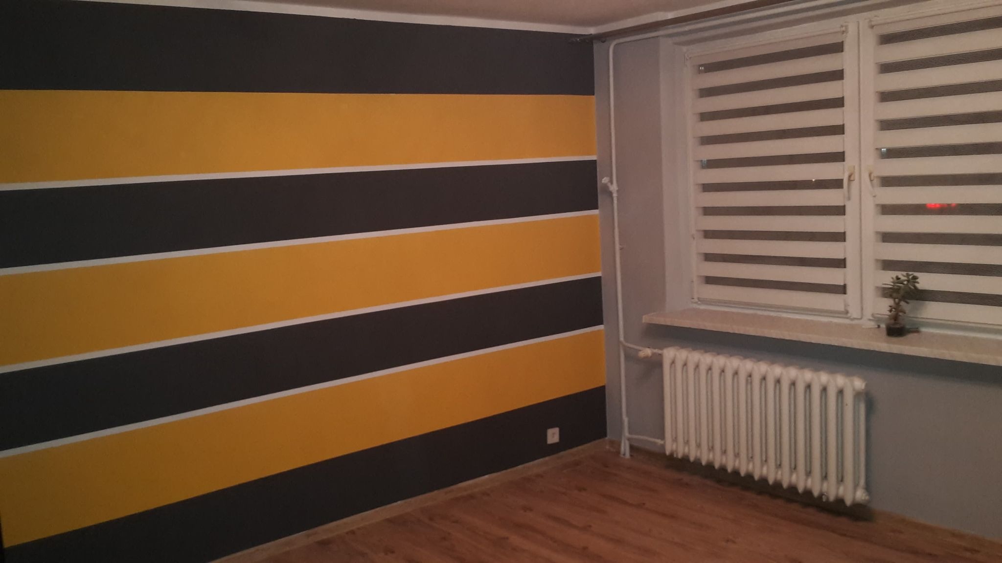 Ściana w pokoju pomalowana w poziome pasy koloru żółtego i ciemnoszarego, z białym paskiem oddzielającym kolory. W tle okno z żaluzjami i grzejnik.