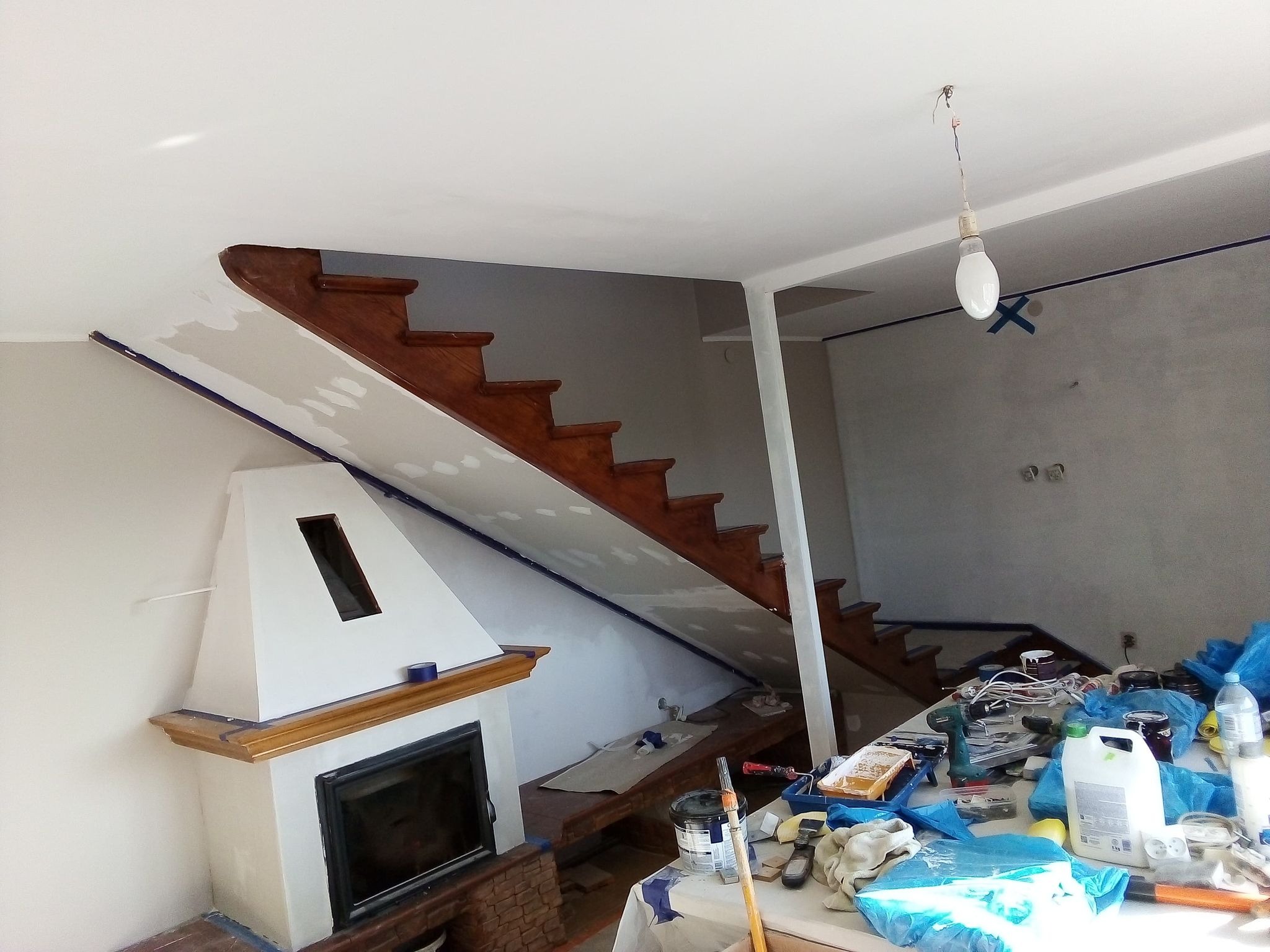 Przygotowania do prac remontowych w salonie: zabezpieczone elementy taśmą malarską, zakryte meble, widoczne narzędzia i materiały malarskie, schody drewniane, kominek z ceglaną podstawą.