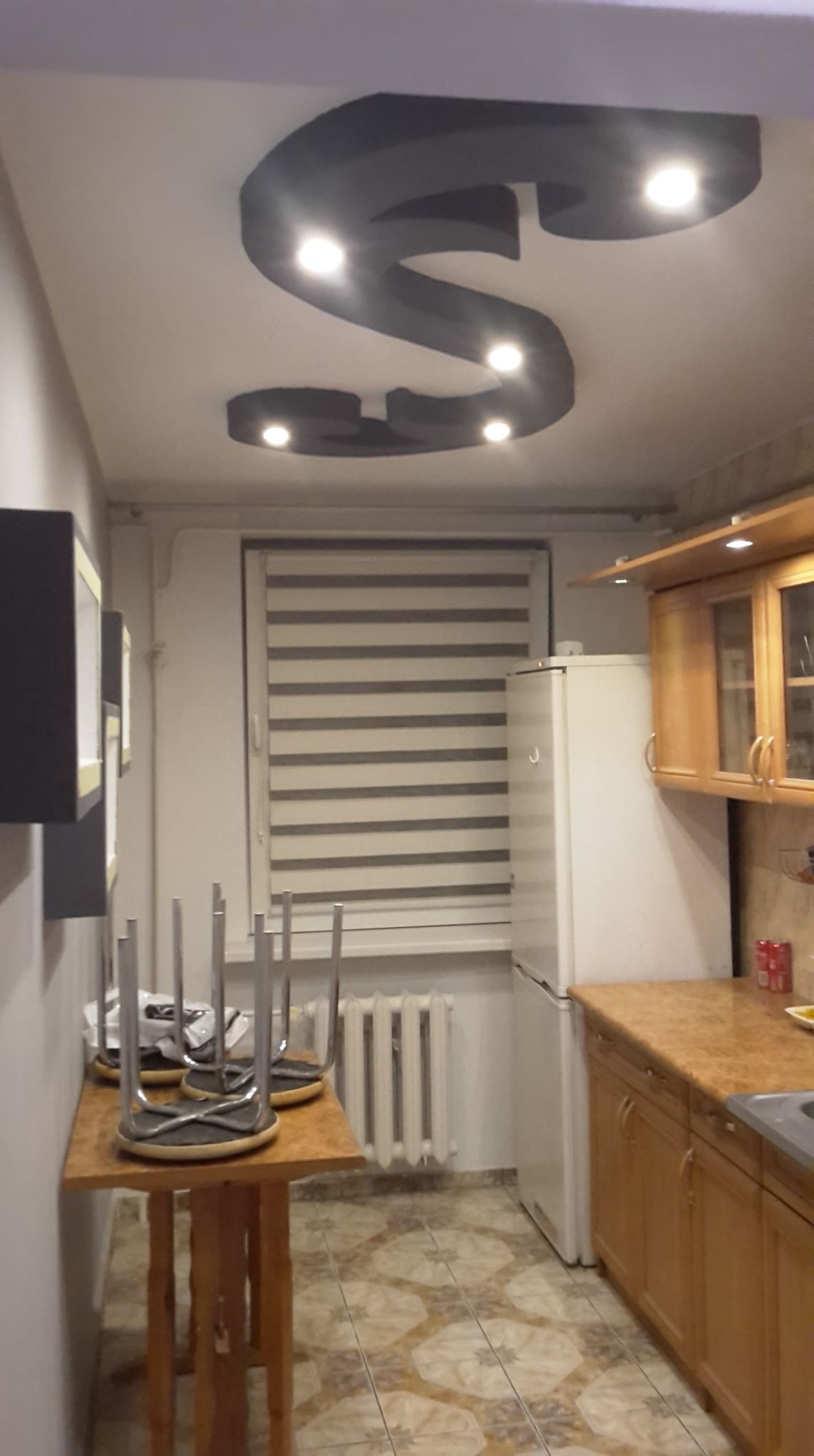 Kuchnia po remoncie z oryginalnym oświetleniem sufitowym w kształcie litery S, widoczne meble kuchenne, okno z roletami i ustawione na stole krzesła.