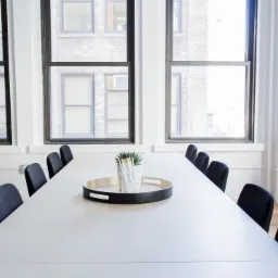 Jasne, minimalistyczne wnętrze sali konferencyjnej z dużym stołem, czarnymi krzesłami i widokiem na budynek za oknem.