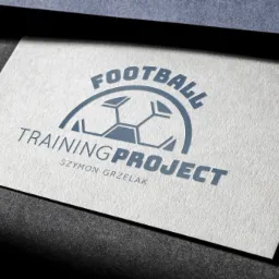 Biała wizytówka z wytłoczonym na szaro-niebiesko logo piłkarskim i nazwą firmy 'Football Training Project Szymon Grzelak', leżąca na ciemnoszarym tle, oświetlona z góry.