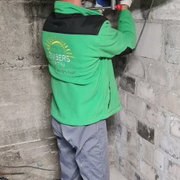 Embers Family Sp. z o.o. - Pracownik w zielonej bluzie z logo Embers Family wierci otwór w ścianie z cegły, używając wiertnicy z odsysaniem pyłu, widoczne rury instalacyjne pod sufitem.