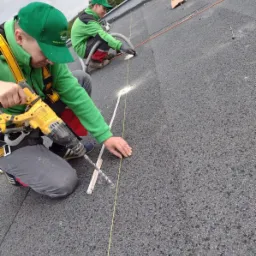 Dwóch monterów w zielonych uniformach, zabezpieczonych uprzężą, instaluje system mocowania na dachu pokrytym papą. Jeden wierci wiertarką DeWalt, drugi odkurza pył wzdłuż wyznaczonej linii.