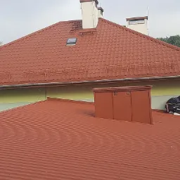 Nowy, czerwony dach z blachy trapezowej, widoczne kominy, okno dachowe i elementy systemu odgromowego. W tle budynek z elewacją w odcieniach zieleni i żółci.