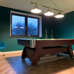 Pok&oacute;j z ciemnozielonymi ścianami i drewnianą podłogą w jodełkę, oświetlony lampą nad stołem bilardowym z brązowego drewna, z widokiem na drzewa za oknem i turkusowym krzesłem na k&oacute;łkach z boku.