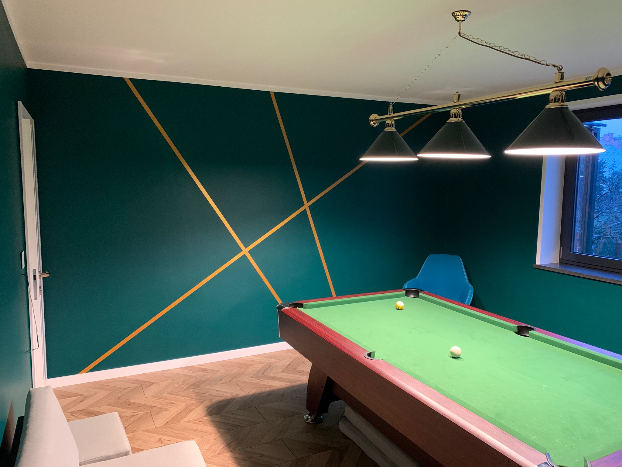 Pokój z zielonymi ścianami, ozdobiony złotymi geometrycznymi liniami, z drewnianym parkietem, stołem bilardowym i lampą wiszącą z trzema kloszami.