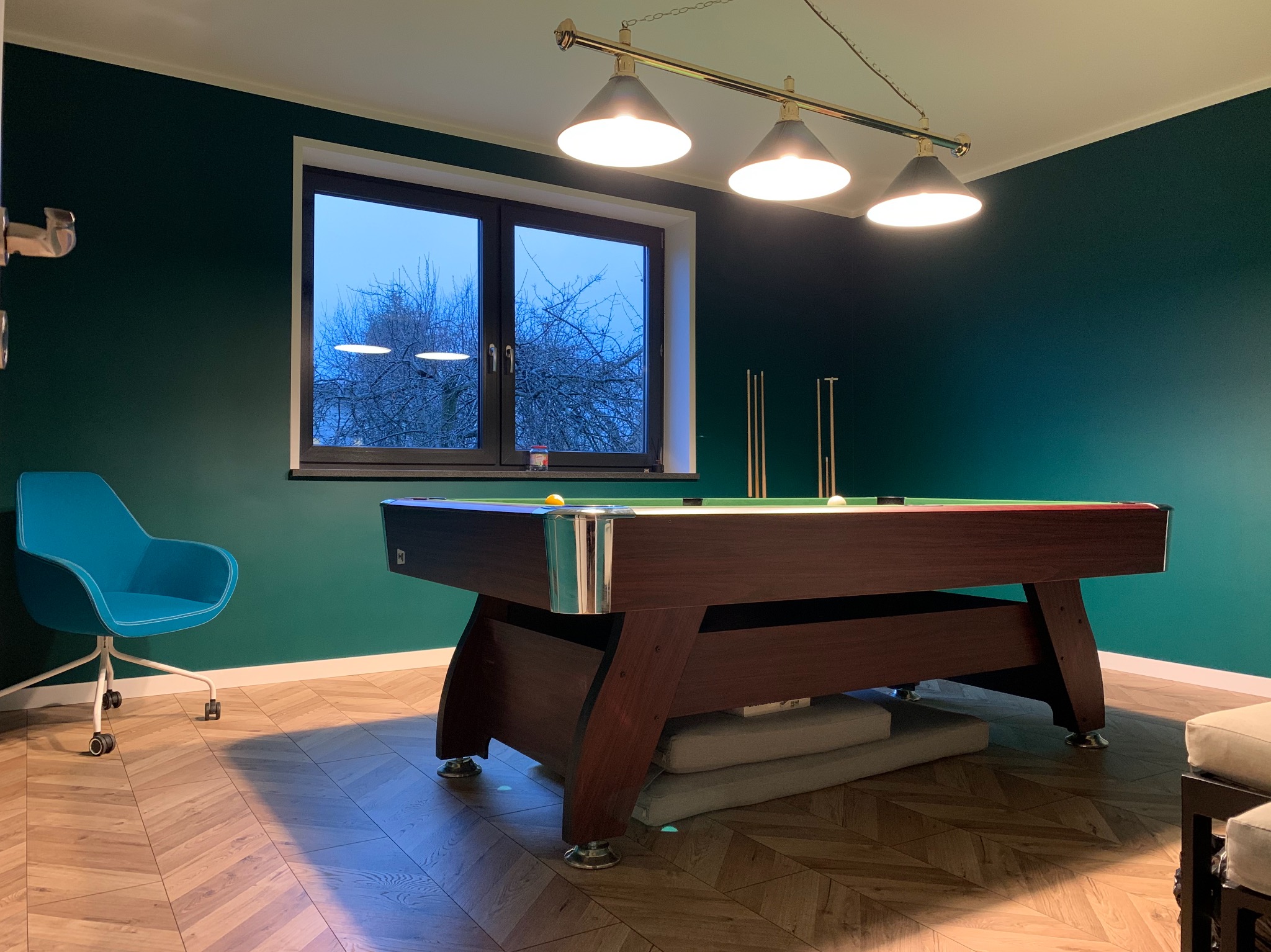 Pokój z ciemnozielonymi ścianami i drewnianą podłogą w jodełkę, oświetlony lampą nad stołem bilardowym z brązowego drewna, z widokiem na drzewa za oknem i turkusowym krzesłem na kółkach z boku.
